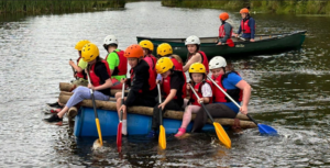 A group of young people row on a lake in a homemade canoe.