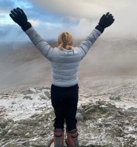 A young person stands top of a peak with their back to the camera. They raise their hands in the air.