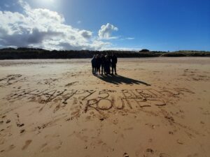 A group of young people stand on a beach. 'Happy 5th birhday Routes,' is written in the sand in front of them.